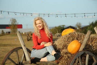 Beautiful woman sitting on wooden cart with pumpkins and hay in field. Autumn season