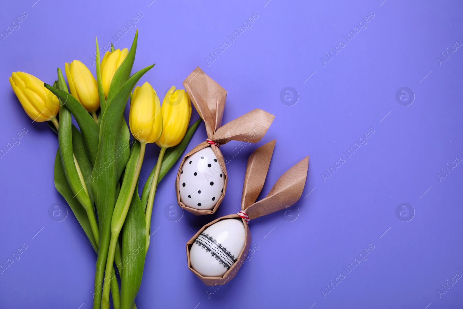 Photo of Easter bunnies made of craft paper and eggs near beautiful tulips on purple background, flat lay. Space for text