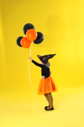 Cute little girl with balloons wearing Halloween costume on yellow background