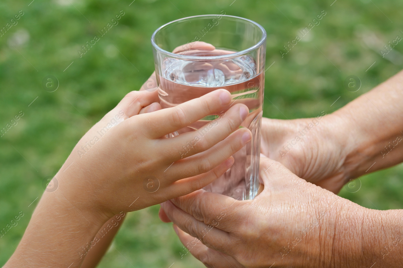 Photo of Child giving glass of water to elderly woman outdoors, closeup