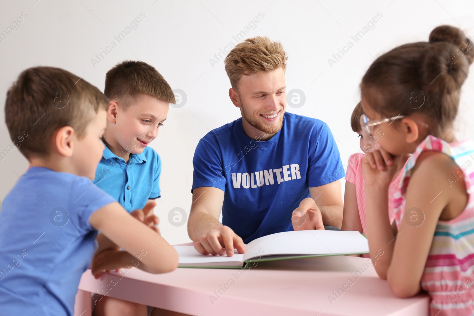 Photo of Young volunteer reading book with children at table indoors