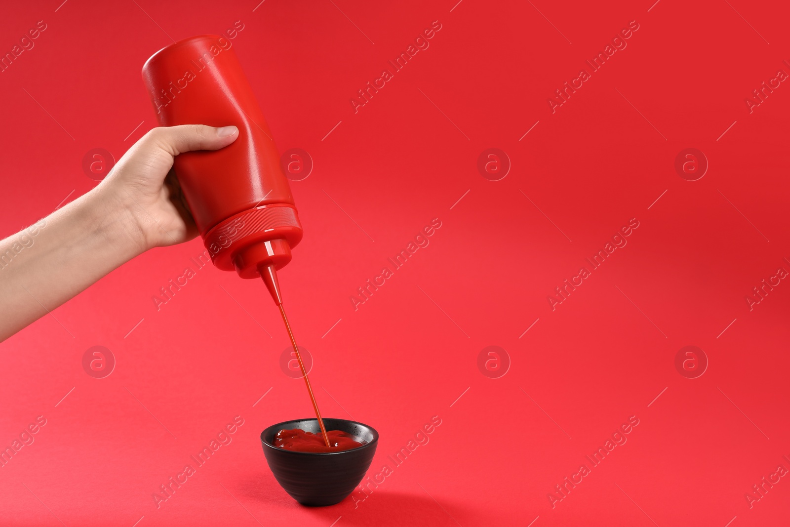 Photo of Woman pouring tasty ketchup from bottle into bowl on red background, closeup. Space for text