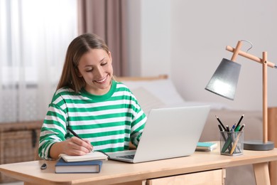 Photo of Online learning. Smiling teenage girl writing in notebook near laptop at home