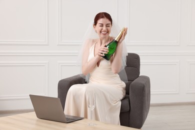 Cheerful bride with bottle of sparkling wine near laptop on wooden table indoors