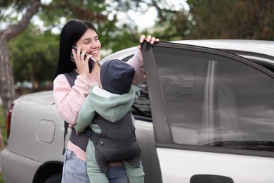 Photo of Mother holding her child in sling (baby carrier) while talking on smartphone near car outdoors