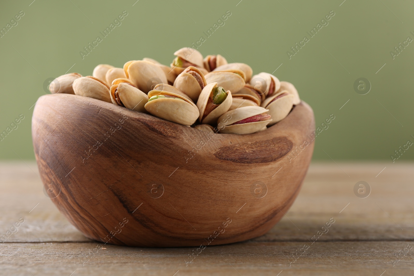 Photo of Tasty pistachios in bowl on wooden table against olive background, closeup