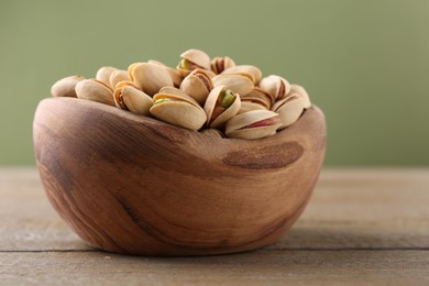 Tasty pistachios in bowl on wooden table against olive background, closeup