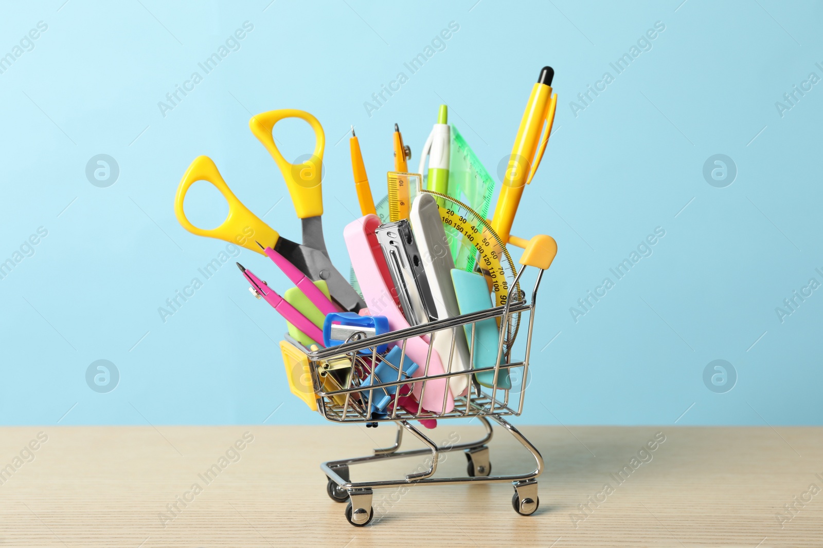 Photo of Small cart with different school stationery on wooden table against light blue background. Back to school