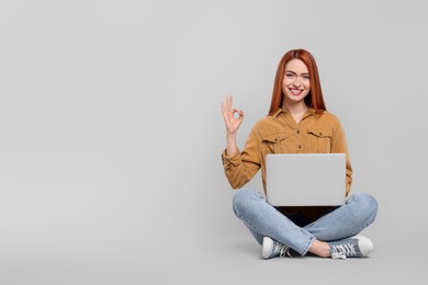 Smiling young woman with laptop showing OK gesture on grey background, space for text