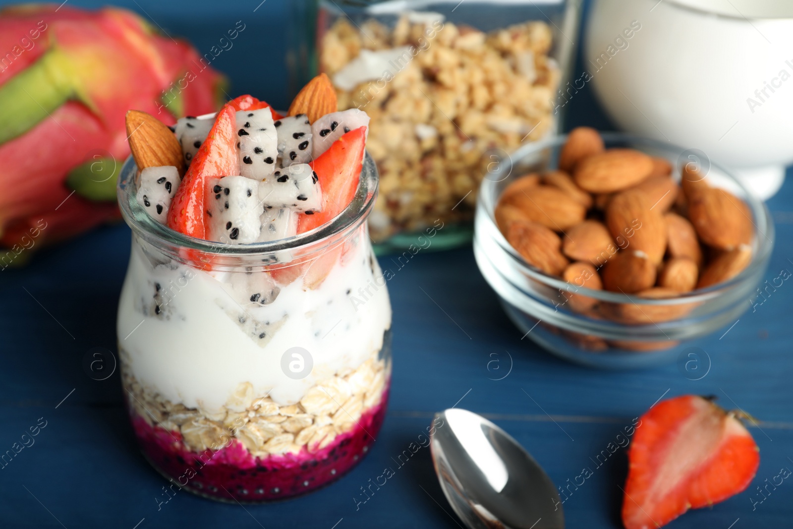 Photo of Glass jar of granola with pitahaya, yogurt and strawberries on blue wooden table, space for text