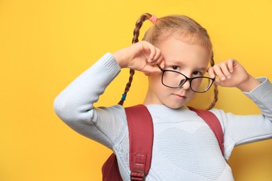Cute little girl with backpack wearing glasses on yellow background