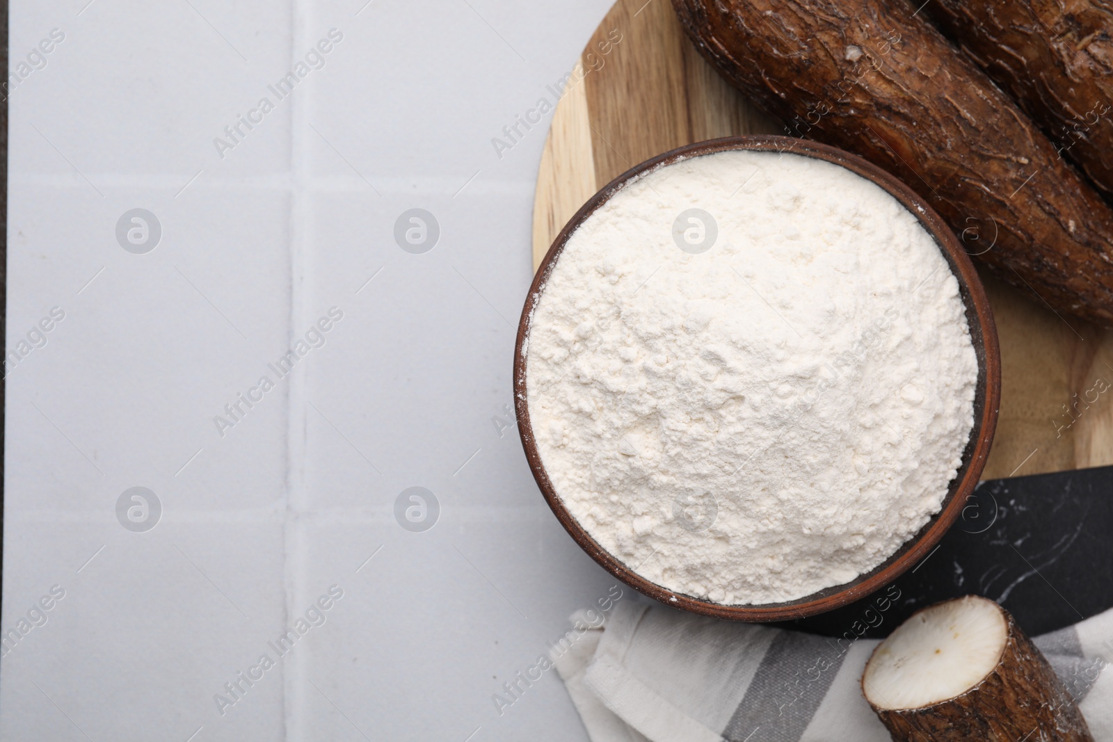 Photo of Bowl with cassava flour and roots on white tiled table, top view. Space for text