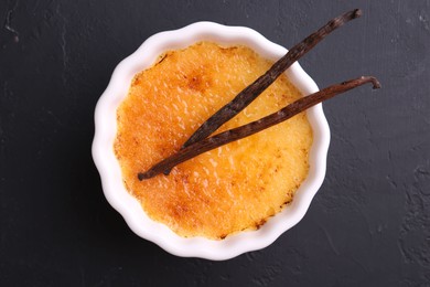 Photo of Delicious creme brulee in bowl and vanilla pods on dark gray textured table, top view