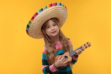 Cute girl in Mexican sombrero hat playing ukulele on orange background