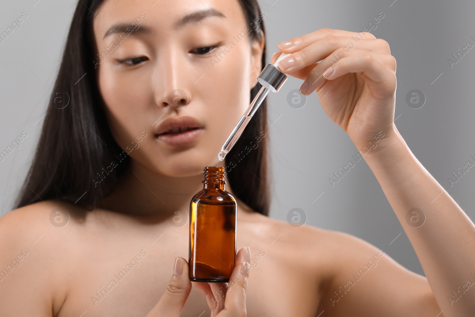 Photo of Beautiful young woman with bottle of cosmetic serum on grey background, selective focus