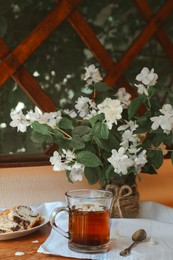 Photo of Glass cup of aromatic tea, tasty dessert and beautiful jasmine flowers on wooden table indoors
