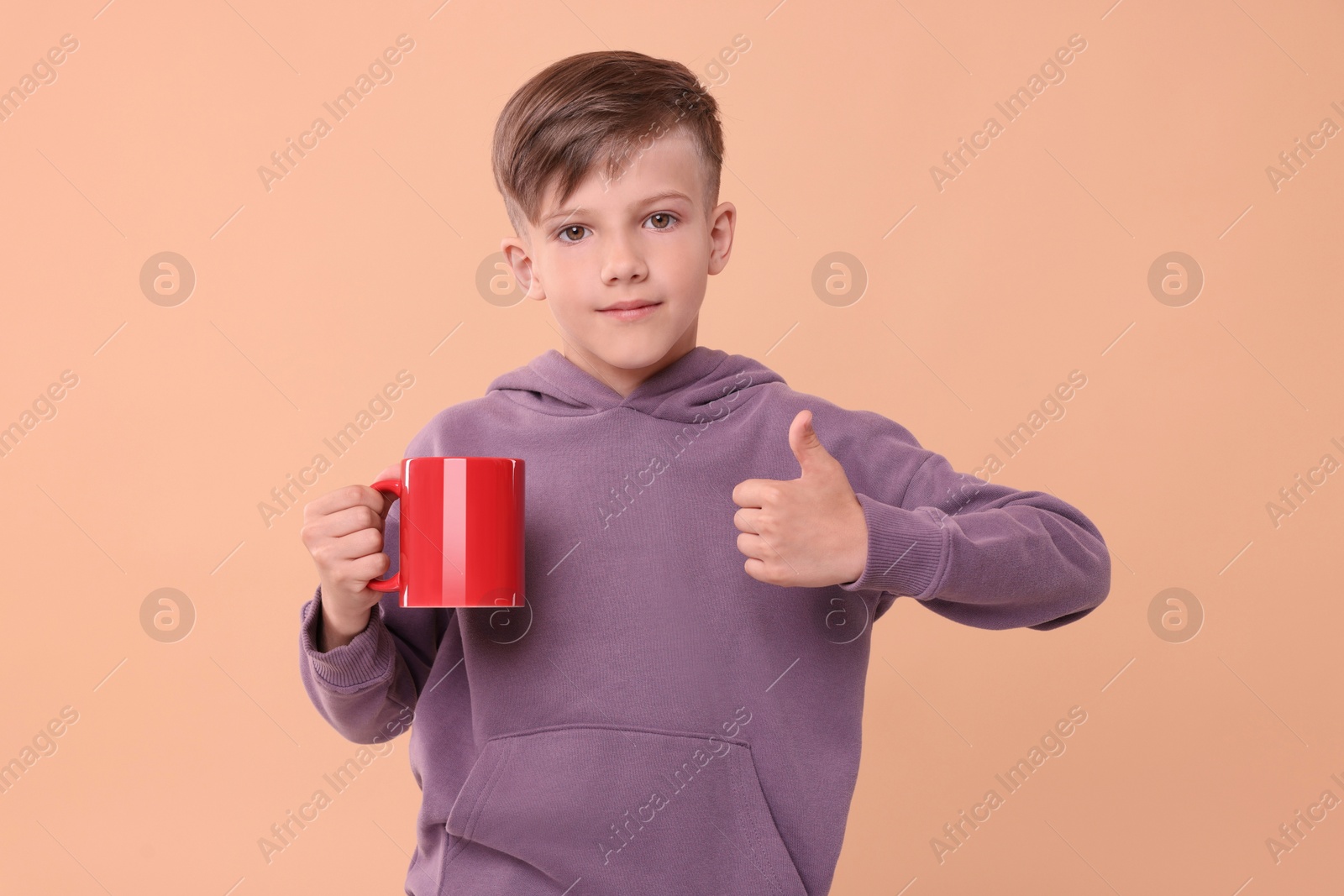 Photo of Cute boy with red ceramic mug showing thumbs up on beige background