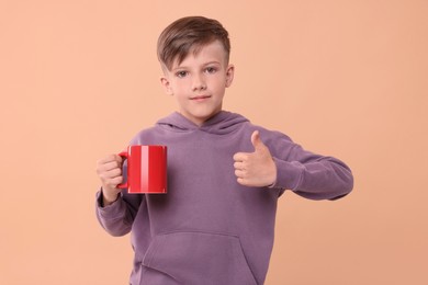 Cute boy with red ceramic mug showing thumbs up on beige background