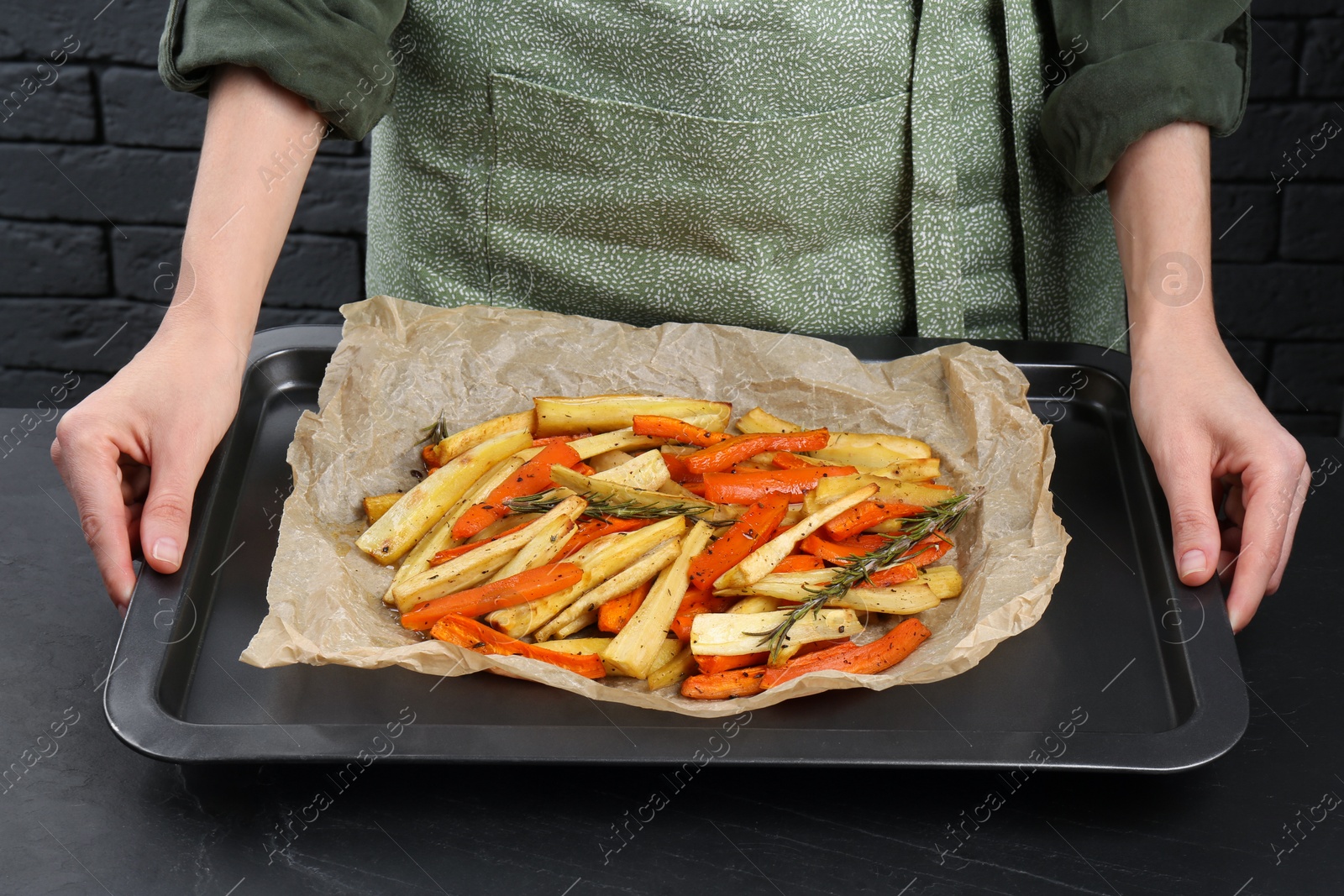 Photo of Cooker holding baking pan with tasty parsnip and bell pepper at black table, closeup