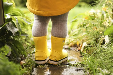 Photo of Little girl wearing rubber boots standing in puddle outdoors, closeup
