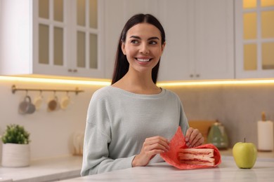 Photo of Happy woman packing sandwich into beeswax food wrap at table in kitchen