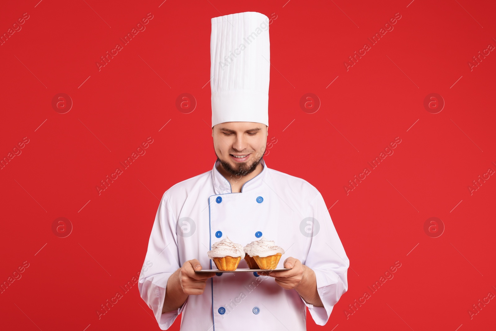 Photo of Happy professional confectioner in uniform holding delicious cupcakes on red background