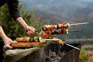 Photo of Woman holding metal skewers and board with delicious meat against mountain landscape, closeup