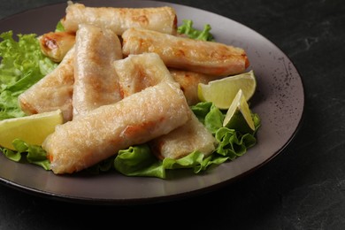 Photo of Plate with tasty fried spring rolls, lettuce and lime on dark textured table, closeup