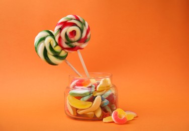 Tasty jelly candies and lollipops in jar on orange background