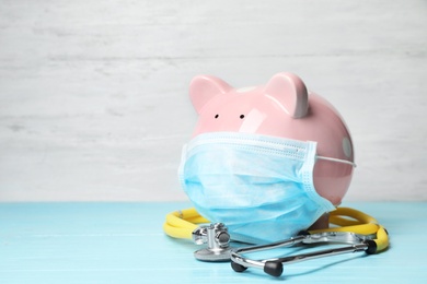 Piggy bank with face mask and stethoscope on table. Space for text