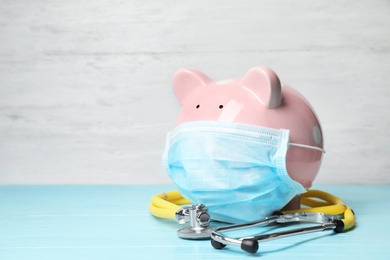 Photo of Piggy bank with face mask and stethoscope on table. Space for text