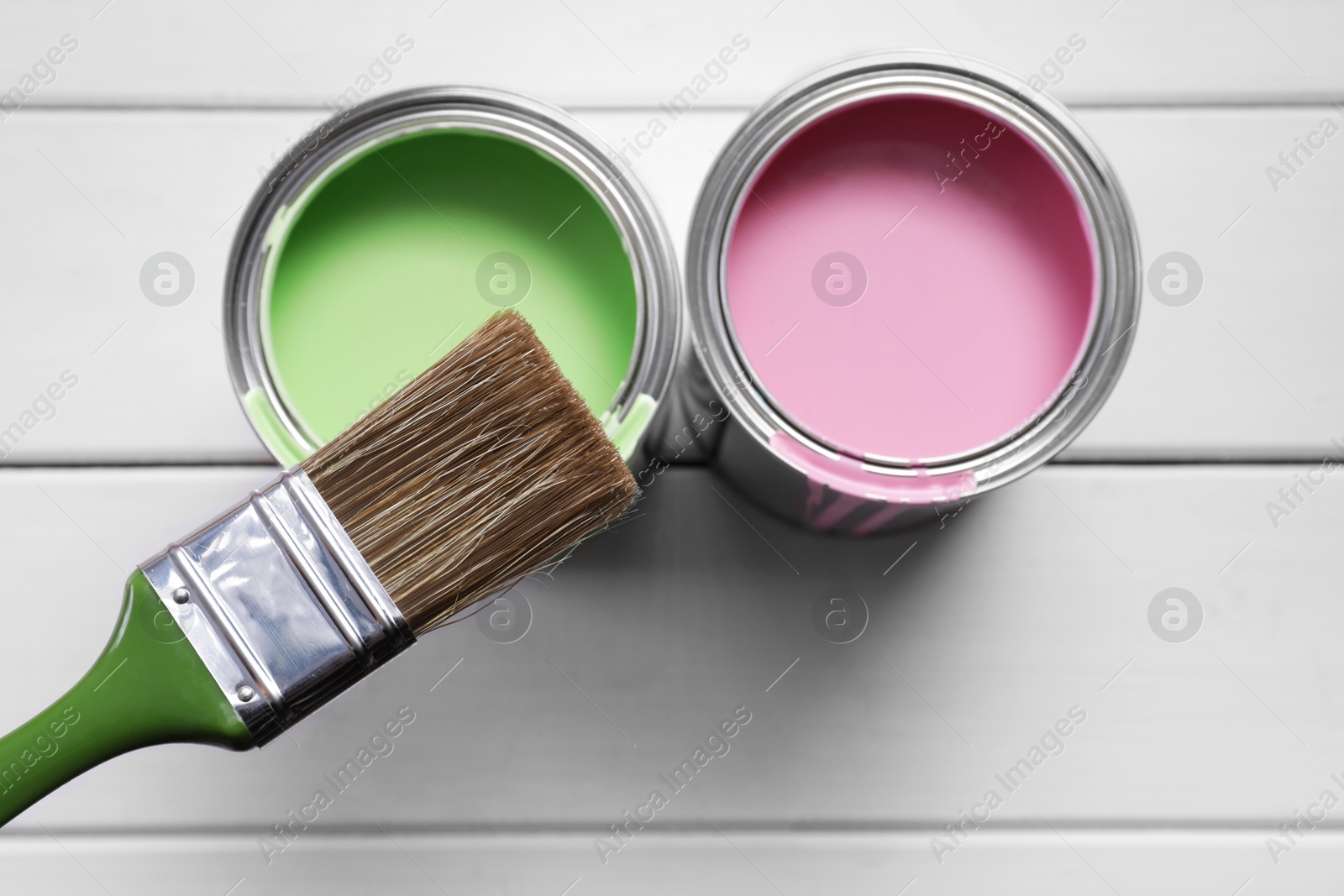 Photo of Cans of pink and green paints with brush on white wooden table, flat lay