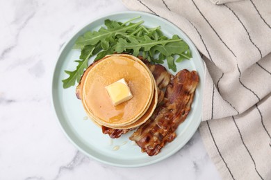 Photo of Tasty pancakes with butter, fried bacon and fresh arugula on white marble table, top view