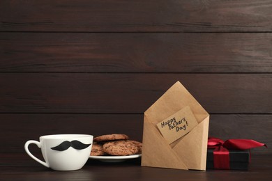 Photo of Card with phrase Happy Father's Day in envelope, cookies, cup with moustache and gift box on wooden table. Space for text