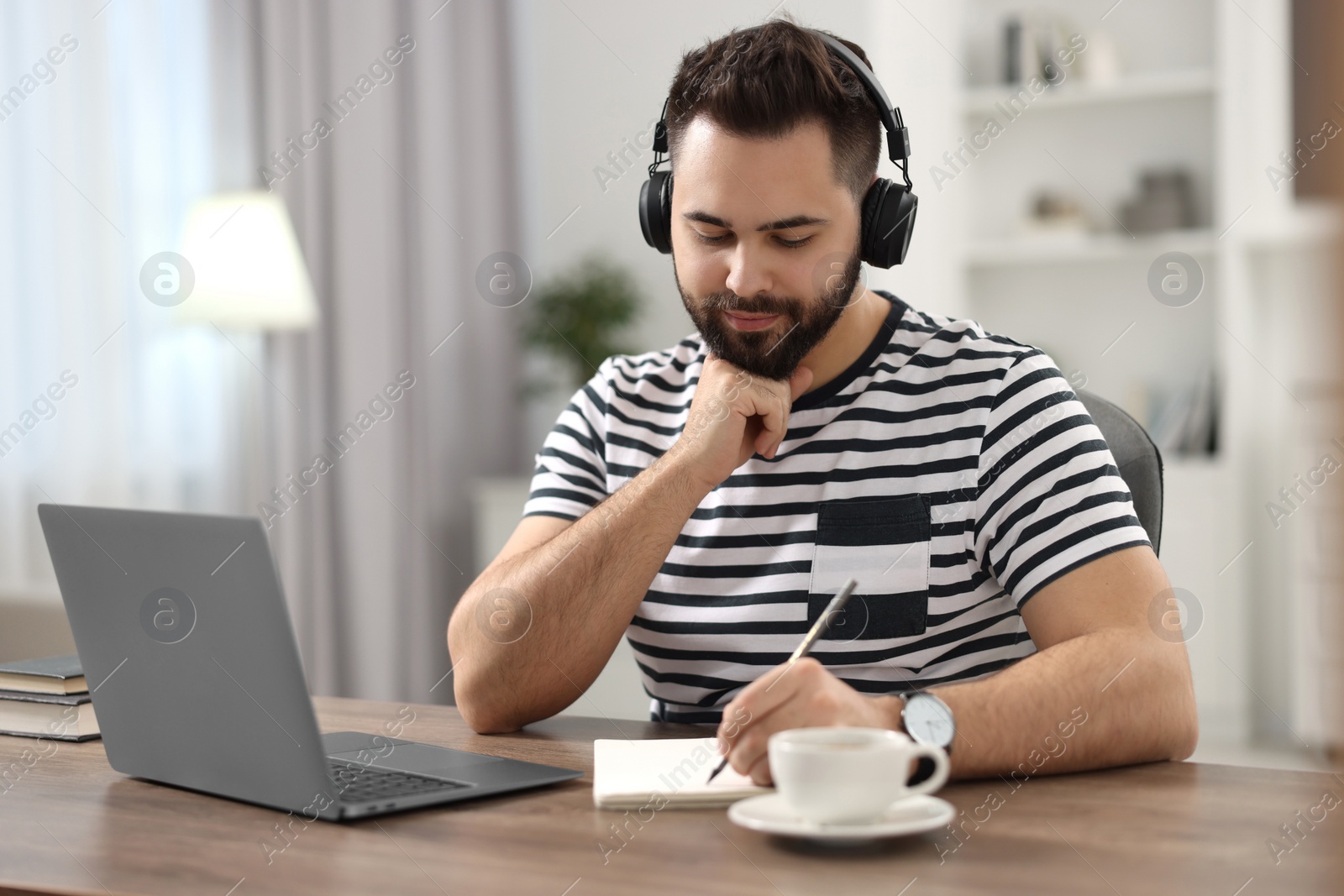 Photo of Young man in headphones writing down notes during webinar at table in room