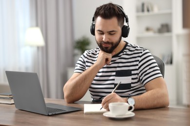 Photo of Young man in headphones writing down notes during webinar at table in room