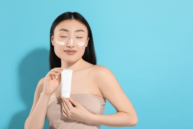 Photo of Beautiful young woman with sunscreen on her face holding sun protection cream against light blue background, space for text