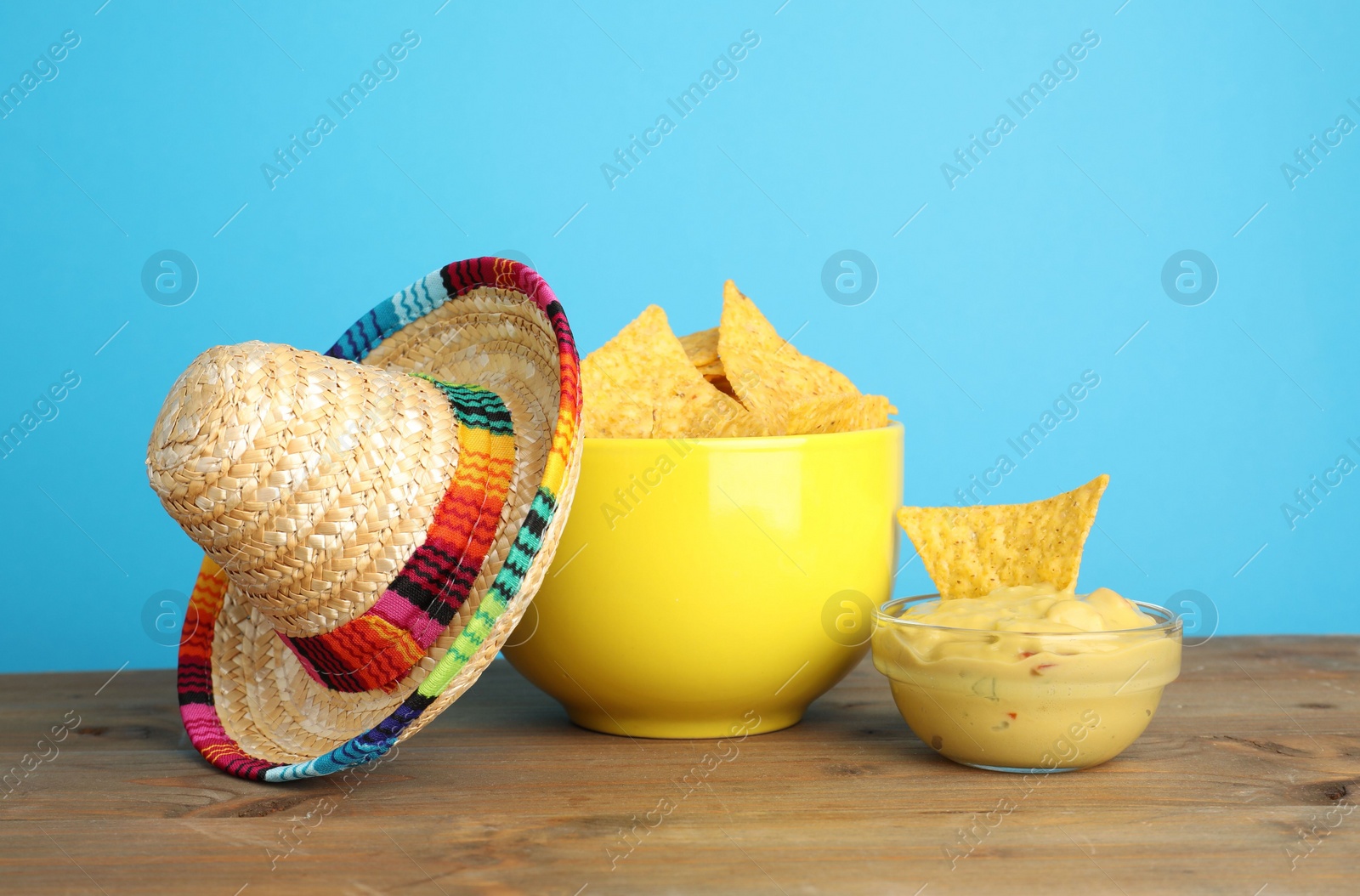 Photo of Mexican sombrero hat, nachos chips and guacamole on wooden table against light blue background