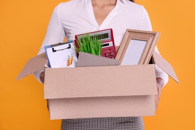 Unemployed woman with box of personal office belongings on orange background, closeup