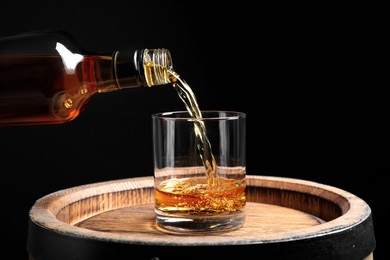 Photo of Pouring whiskey from bottle into glass on wooden barrel against black background, closeup