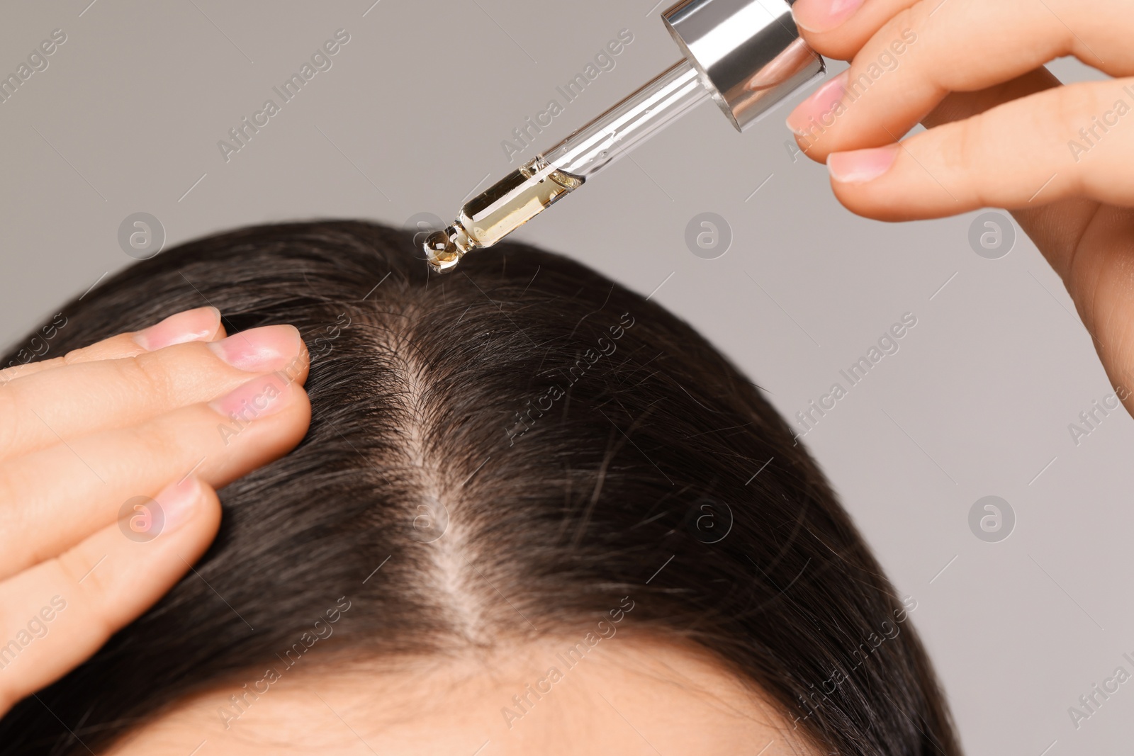 Photo of Woman applying essential oil onto hair roots on light grey background, closeup