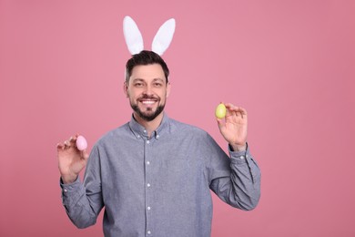Happy man in bunny ears headband holding painted Easter eggs on pink background