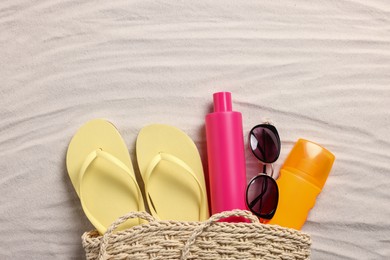 Wicker beach bag with sunscreen, flip flops and sunglasses on sand, flat lay