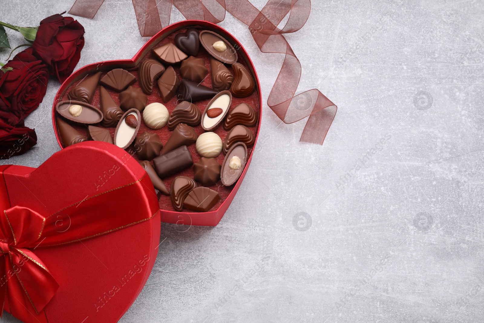 Photo of Heart shaped box with delicious chocolate candies, roses and ribbon on light grey table, flat lay. Space for text