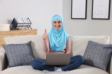 Photo of Muslim woman showing thumb up while using laptop at couch in room