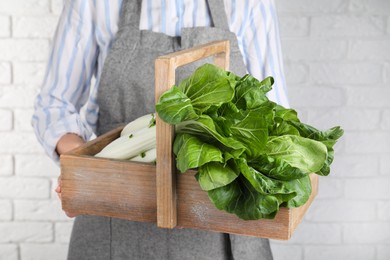 Photo of Woman holding wooden crate with bok choy cabbage near white brick wall, closeup