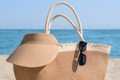 Photo of Stylish straw bag with visor cap and sunglasses on beach near sea, closeup