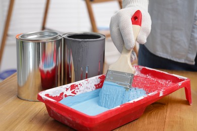 Photo of Man taking light blue paint with brush from tray at wooden table indoors, closeup
