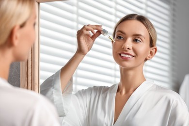 Photo of Beautiful woman applying cosmetic serum onto her face near mirror in bathroom