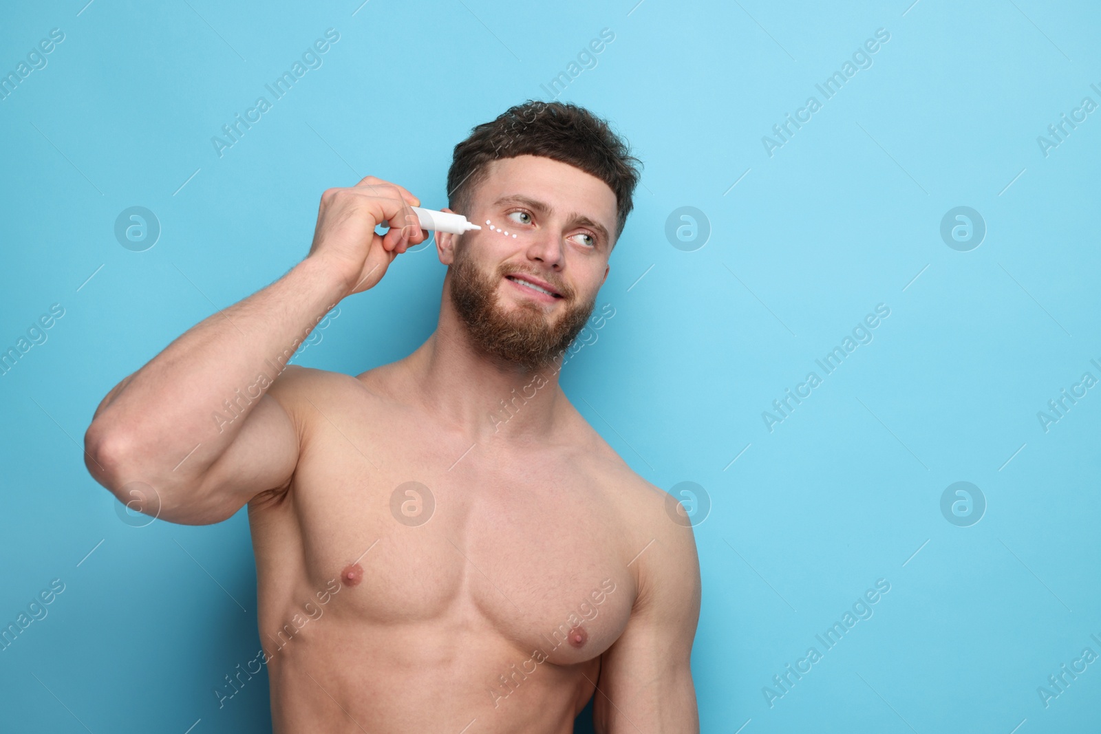 Photo of Handsome man applying moisturizing cream onto his face on light blue background, space for text